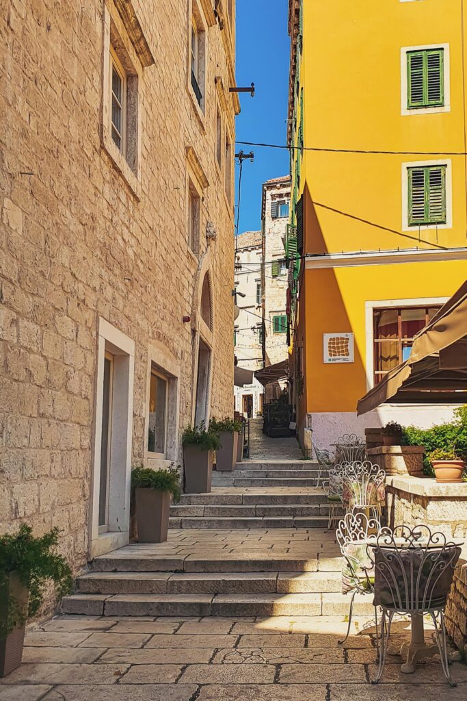 Šibenik yellow house Sunlit narrow alley between stone buildings, with a bright yellow wall and green shutters, and wrought-iron chairs along stone steps, views to admire on a Šibenik day trip