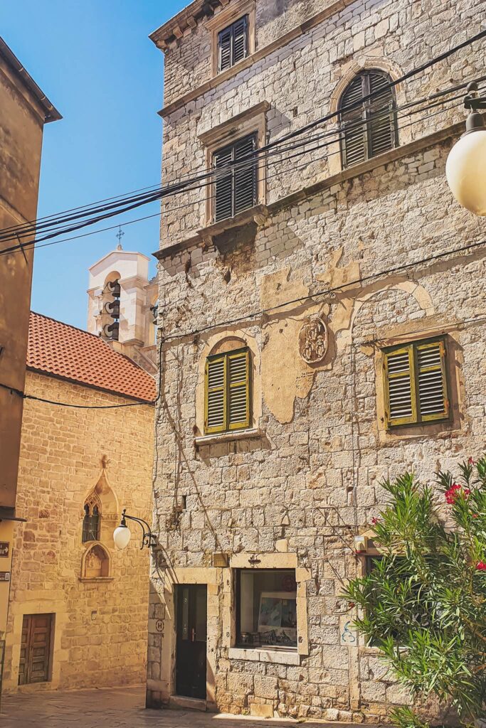 Šibenik stone houses Stone building with arched window and green shutters on a sunny street; a church bell tower is visible in the distance.