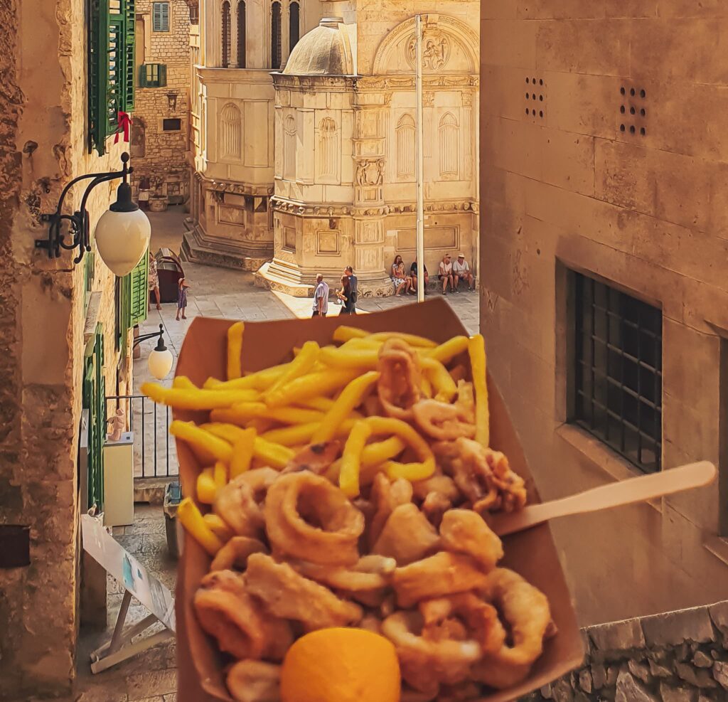 Šibenik fried fish Carton of french fries and fried fish held close to the camera with a sunlit historic street and ornate stone building in the background, and people sitting nearby.