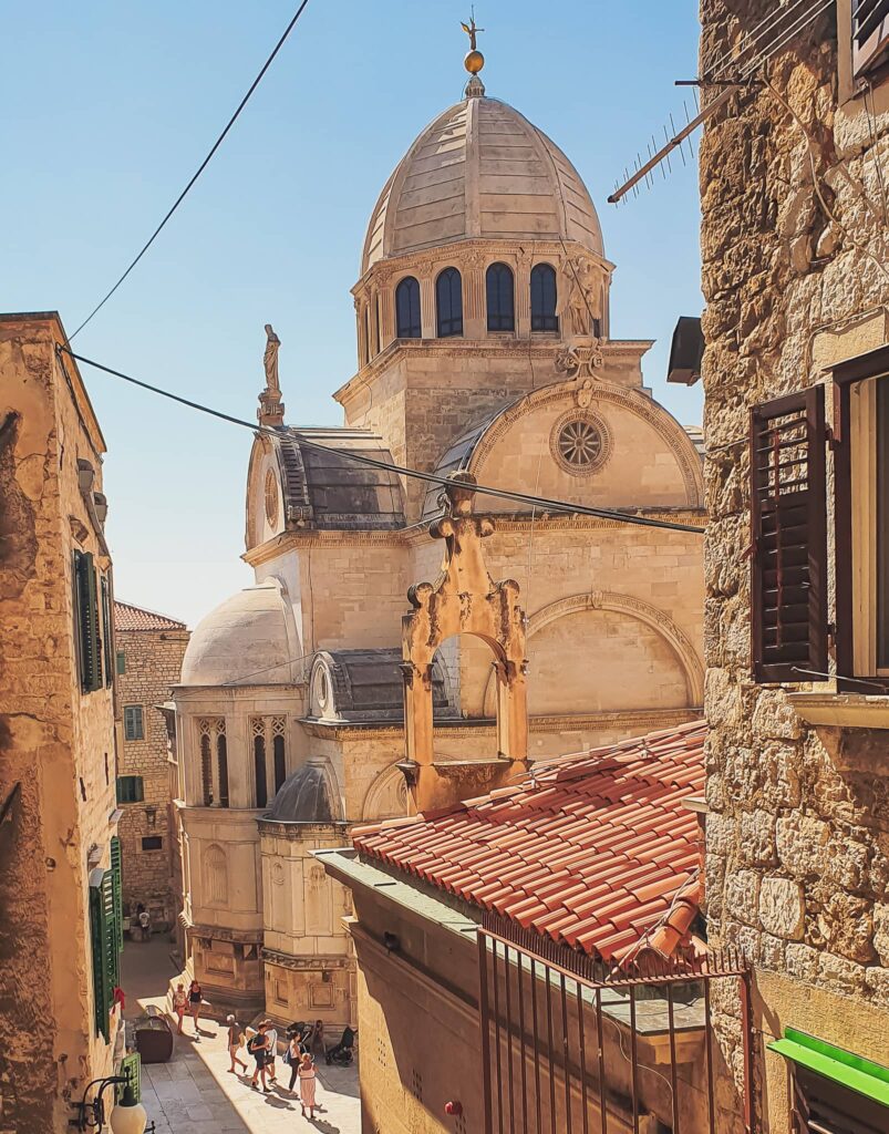 Šibenik Cathedral Stone church with a domed roof and statues, set in a sunlit narrow alley; a group of tourists stroll below.