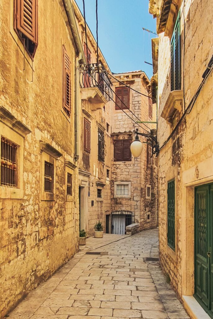 Šibenik Old Town alley Narrow sunlit stone alley in a Mediterranean town, with aged walls, brown wooden shutters, and a cobblestone path.