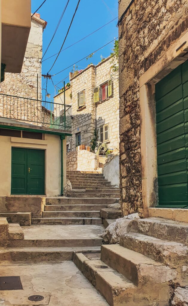 Šibenik stairs Stone stairway between sunlit stone buildings with green doors and shutters, under a clear blue sky.