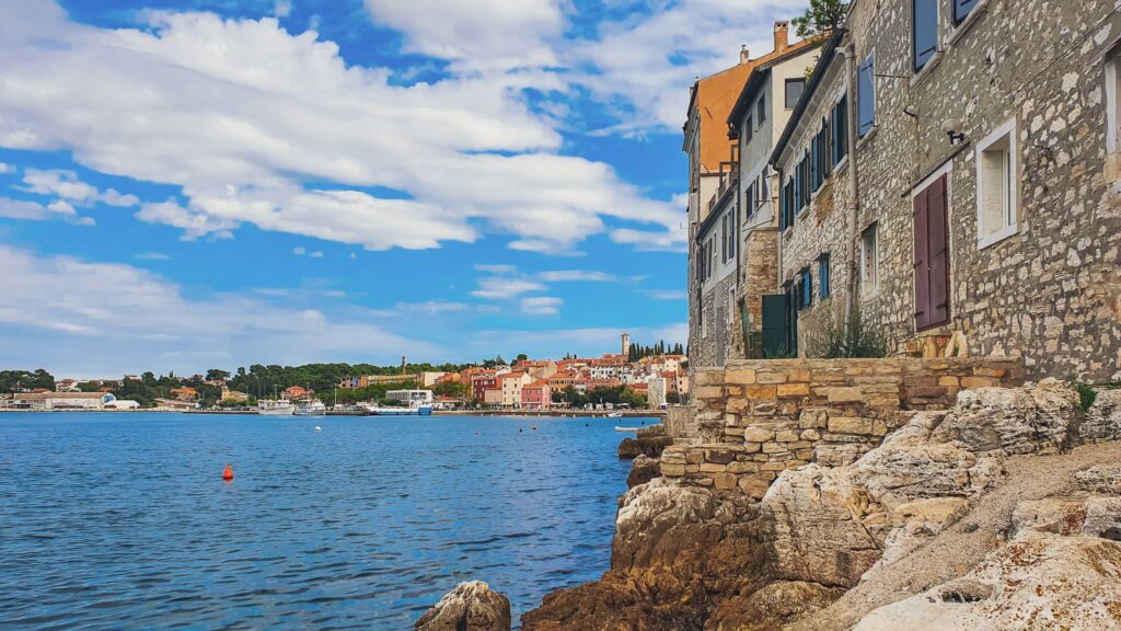 Rovinj coast Stone waterfront building sits on rocky shore overlooking a blue harbor, with a village and boats in the distance under a bright sky.