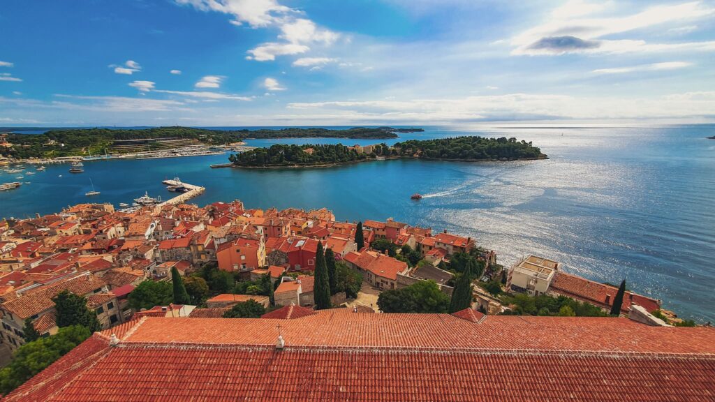 Rovinj and Sveta Katarina Coastal town with red-tiled roofs along the harbor and blue sea, small island in the background.