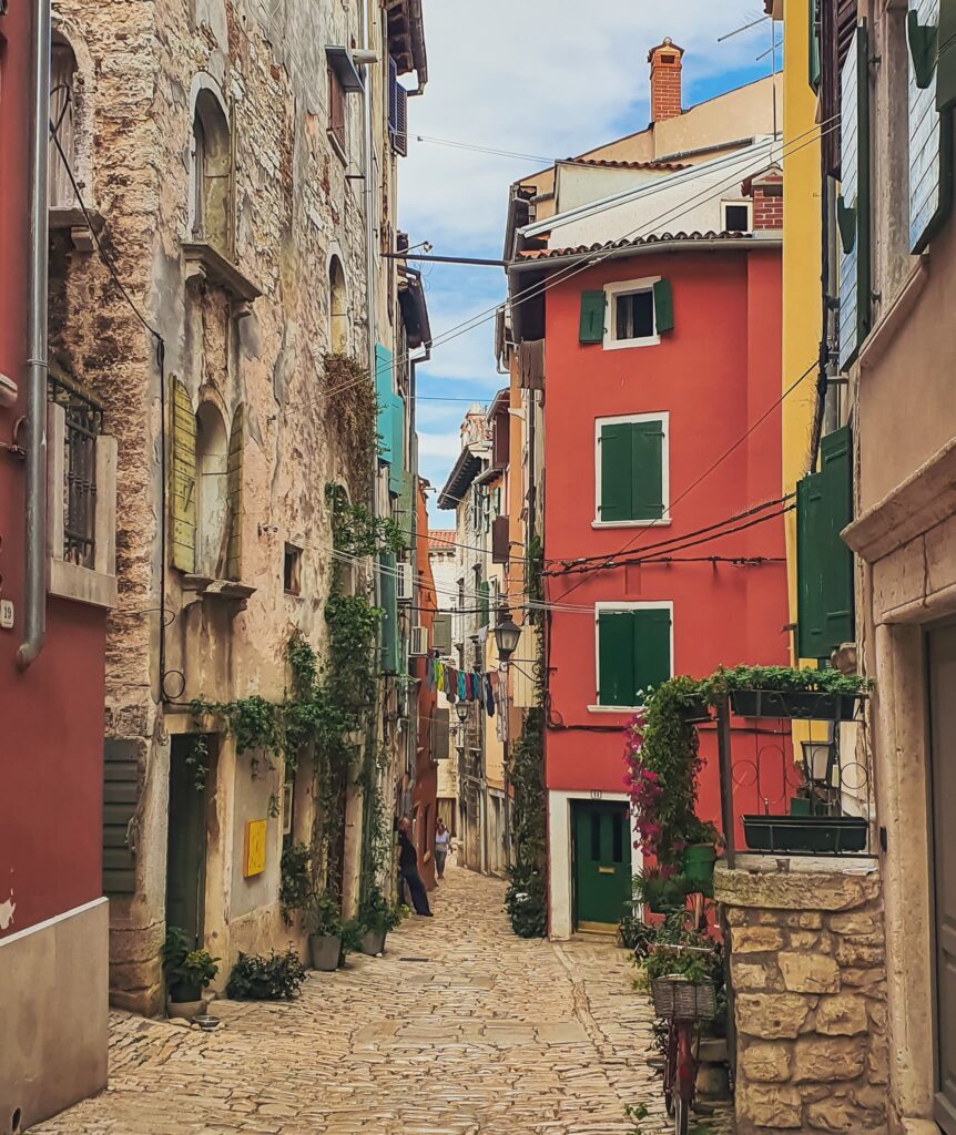 Rovinj Old Town Narrow cobblestone street lined with colorful buildings, green shutters, potted plants, and hanging laundry between stone walls and pastel facades under a blue sky.