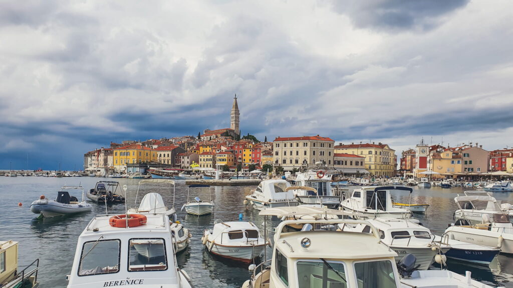 Rovinj Marina Harbor of a coastal town with colorful buildings and a tall church tower, boats docked in the foreground under a cloudy sky